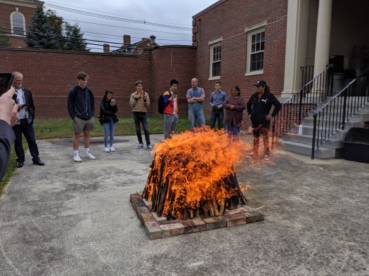 Image of very hot orange fire burning with Native American artists and Phillips Academy students looking on in the background.