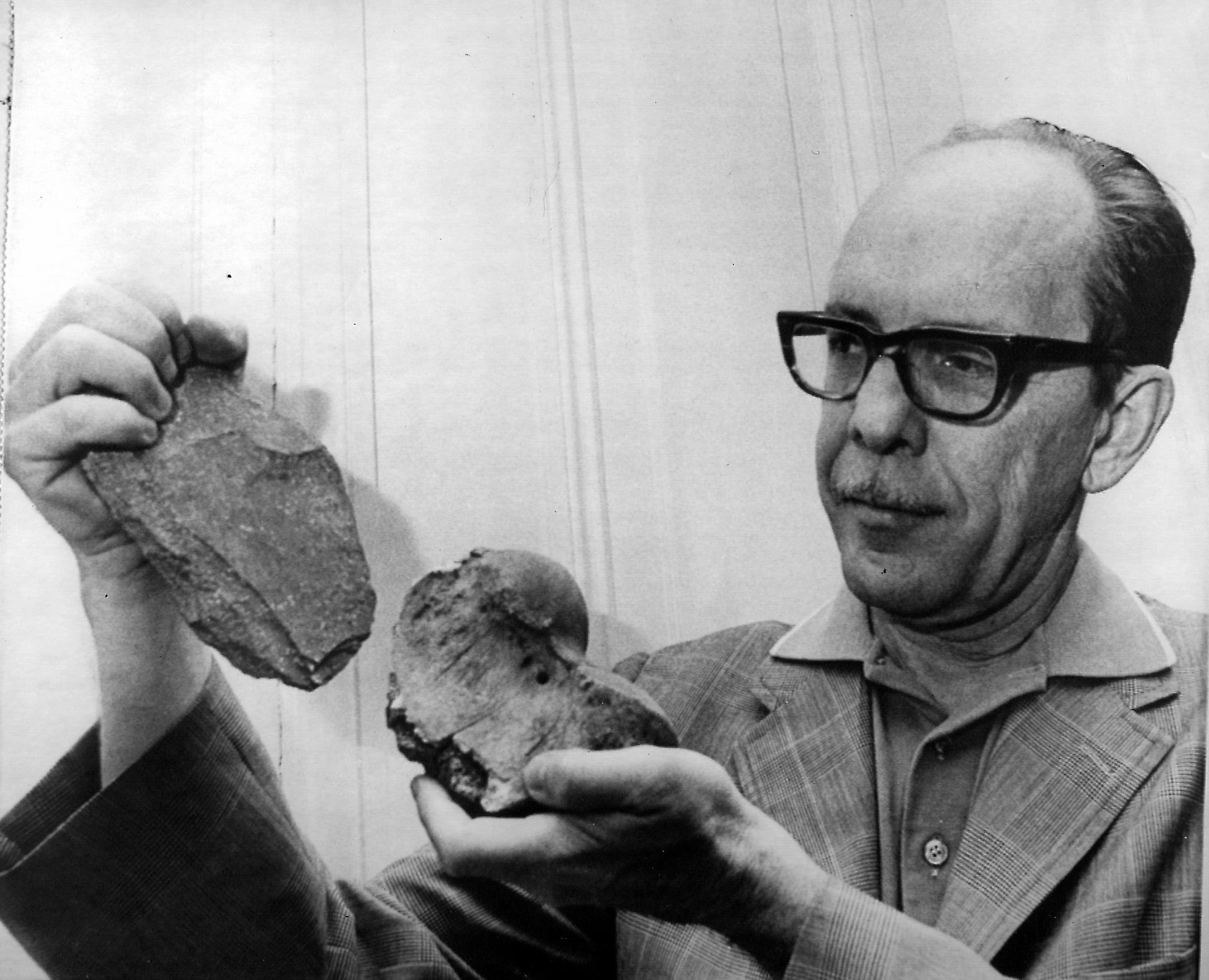Image of Scotty MacNeish, wearing heavy black framed glasses and a tweed blazer holding a large, crude stone chopper tool and the end of a large sloth leg bone.