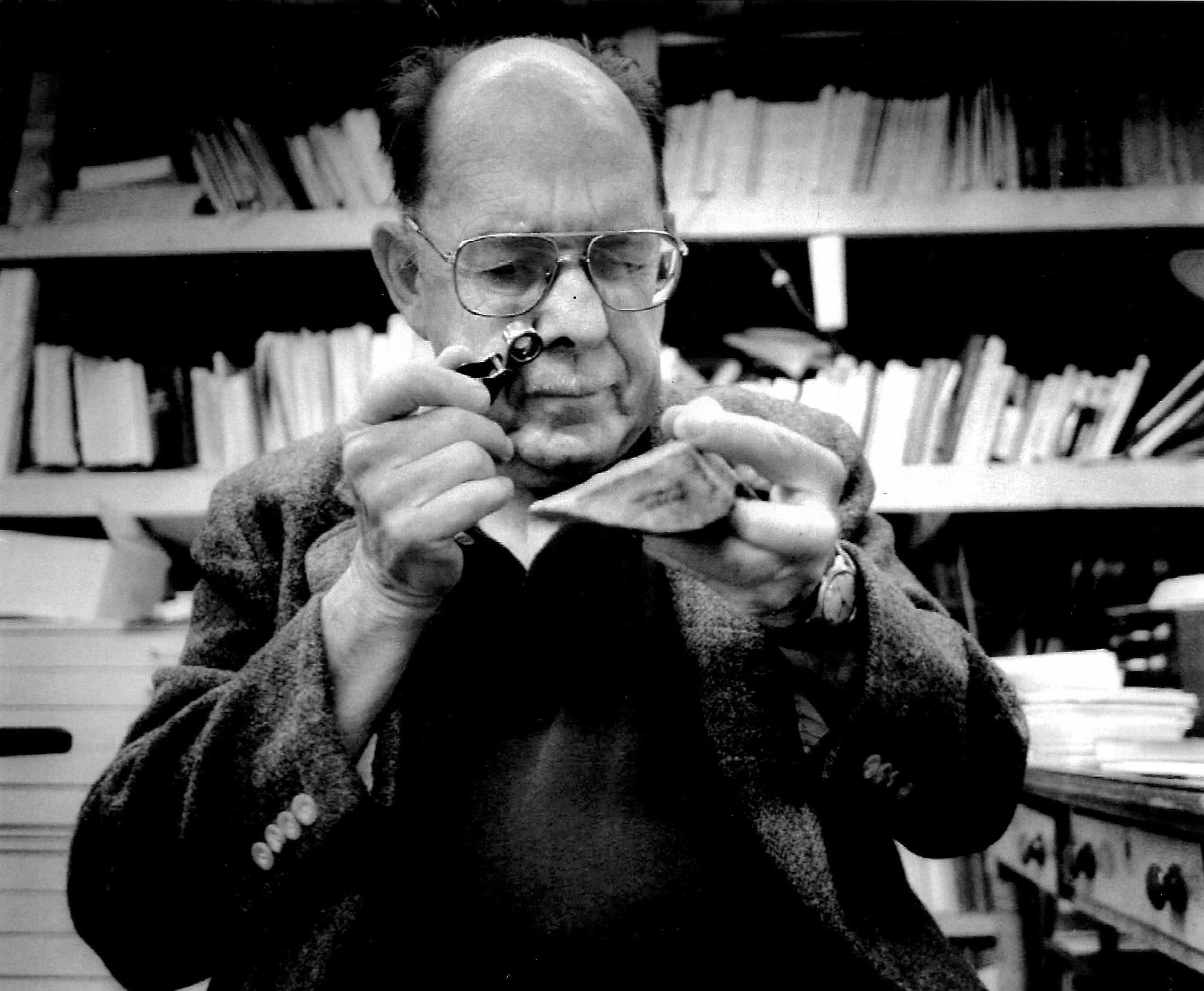 Image of Scotty MacNeish, an older, balding man with wire frame glasses using a jeweler's loupe to examine a point stone tool from Pendejo Cave, New Mexico. Bookshelves are in the background, slightly out of focus.