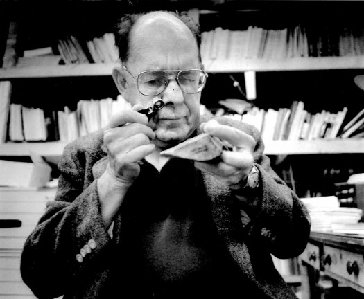 Image of Scotty MacNeish, an older, balding man with wire frame glasses using a jeweler's loupe to examine a point stone tool from Pendejo Cave, New Mexico. Bookshelves are in the background, slightly out of focus.