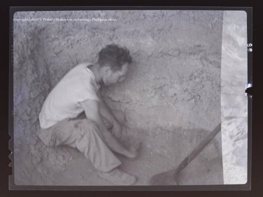 A man in a white t-shirt and khaki pants sits in an excavation pit and removes samples.