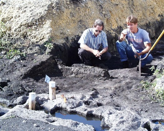 Two male archaeologists crouch down and examine water-filled basins carved in limestone bedrock. Dirt piles are behind them.