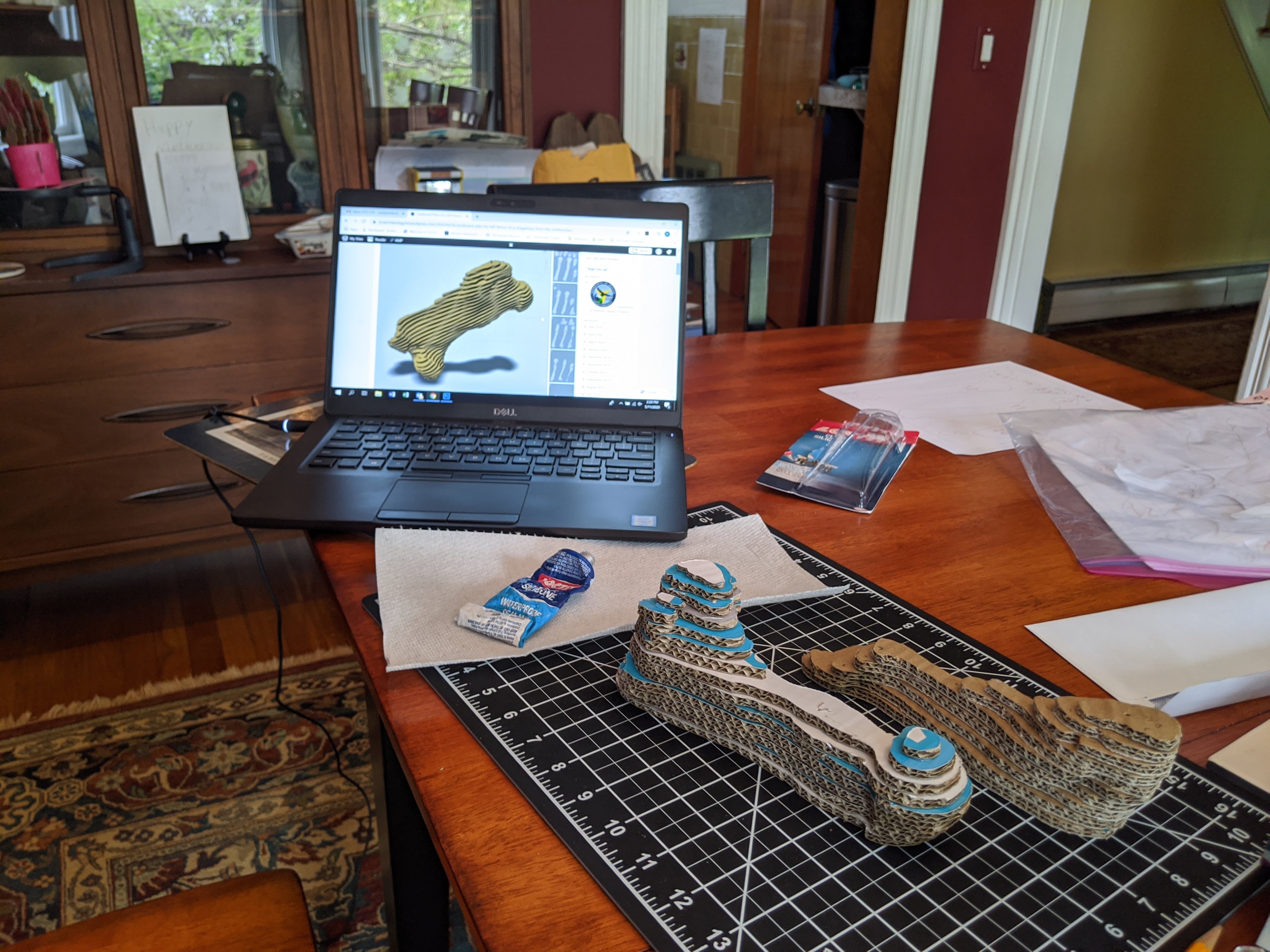Image of stacked cardboard pieces on a cutting mat with a computer in the background showing a virtual assembly of the model.