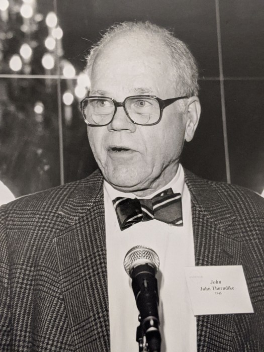 Image of John Thorndike, an older man with black rim glasses, a striped bow tie, and tweed jacket standing in front of a microphone at a museum event.