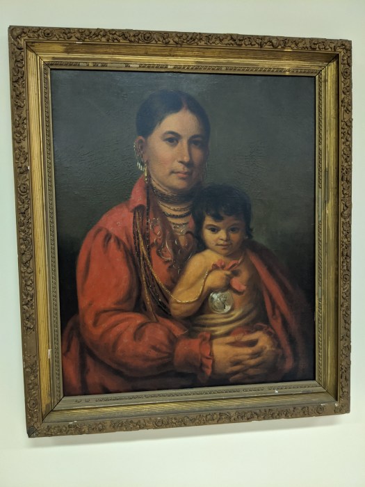 Image shows an oil painting in an elaborate gold gilt frame that depicts an attractive Native American woman holding her baby. She wears a red blouse which covers the baby's shoulders. A silver Indian Peace Medal around her neck is held by the baby.