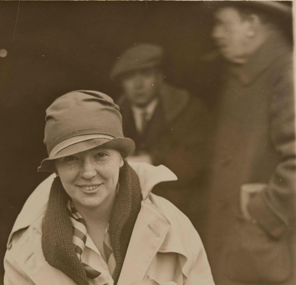 Image of Margaret Ashley as a smiling young woman wearing a cloche hat and light-colored trench coat with collar turned up. She has several scarves loosely around her neck. Hazy, out of focus image of Warren Moorehead in the background.
