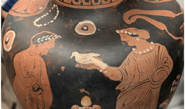 close up image of a painting on a greek vessel, showing a woman holding a dove out toward a man wearing a laurel wreath