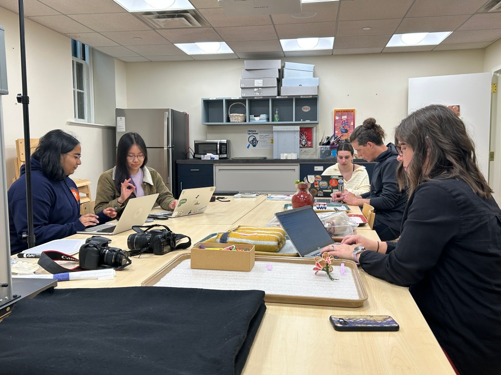 a group of five people sitting around a large table covered in objects, typing on computers