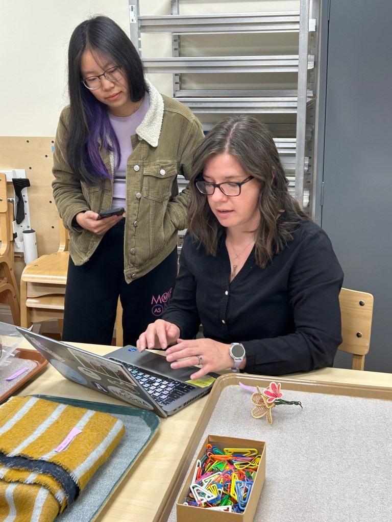 a woman sitting at a table with objects in front of her, typing on her laptop and talking to another woman standing behind her and looking over her shoulder