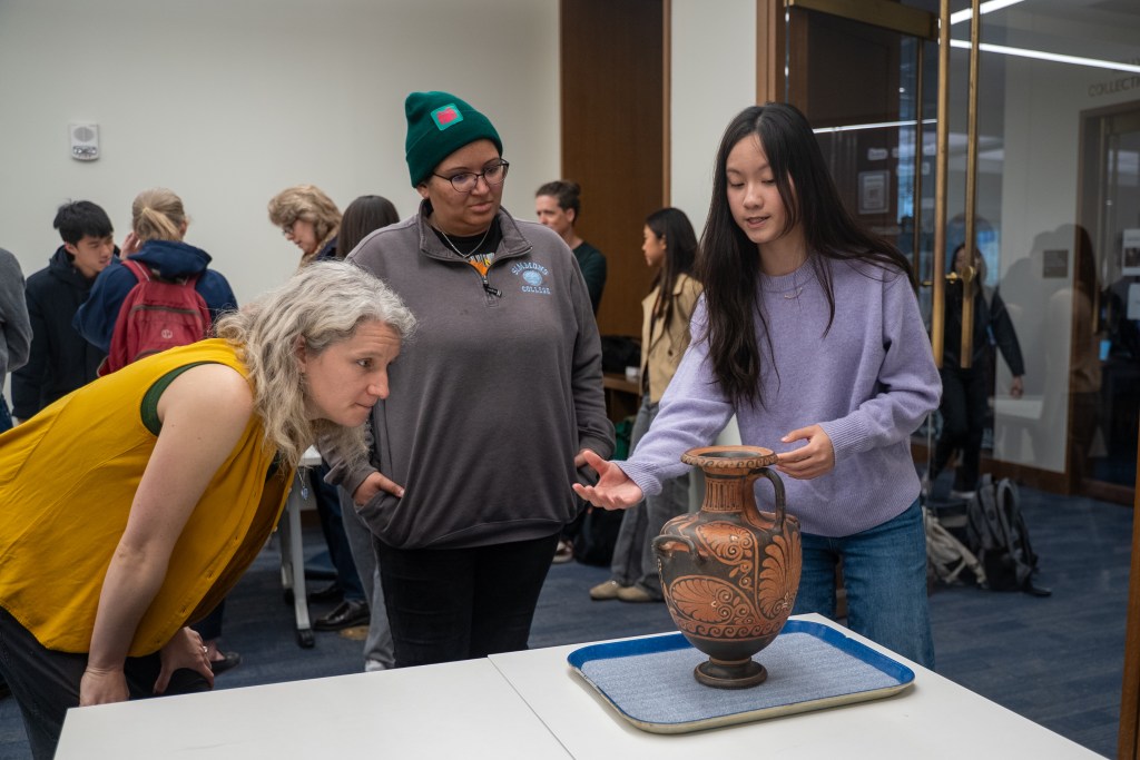 three people standing around an ancient greek pot, looking and discussing