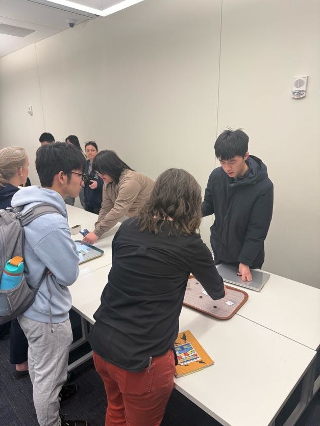 A group of people standing around tables, looking at coins laid out on trays and talking.